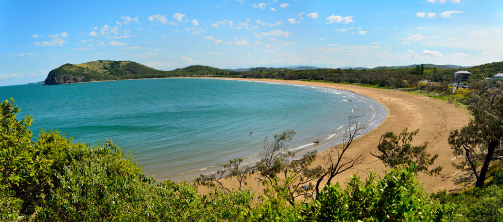 A wide, crescent-shaped sandy beach curving along a calm turquoise bay, bordered by lush green hills and trees under a clear blue sky.
