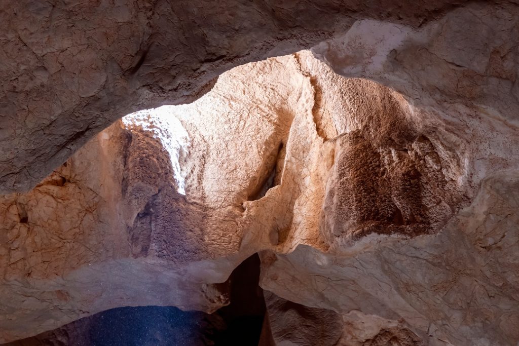An interior view looking up at the rugged, textured rock formations of a cave ceiling, where natural light streams in from an opening above.