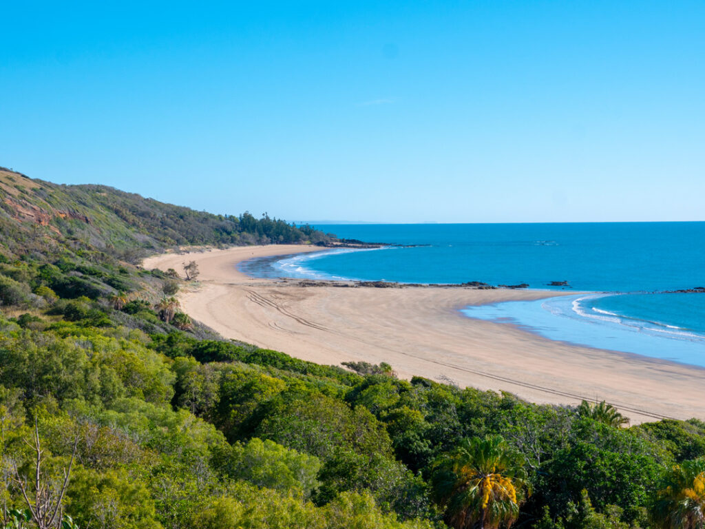 An elevated, wide-angle view of a long, curved sandy beach meeting a calm blue ocean, bordered by lush green coastal vegetation and hills under a clear blue sky.