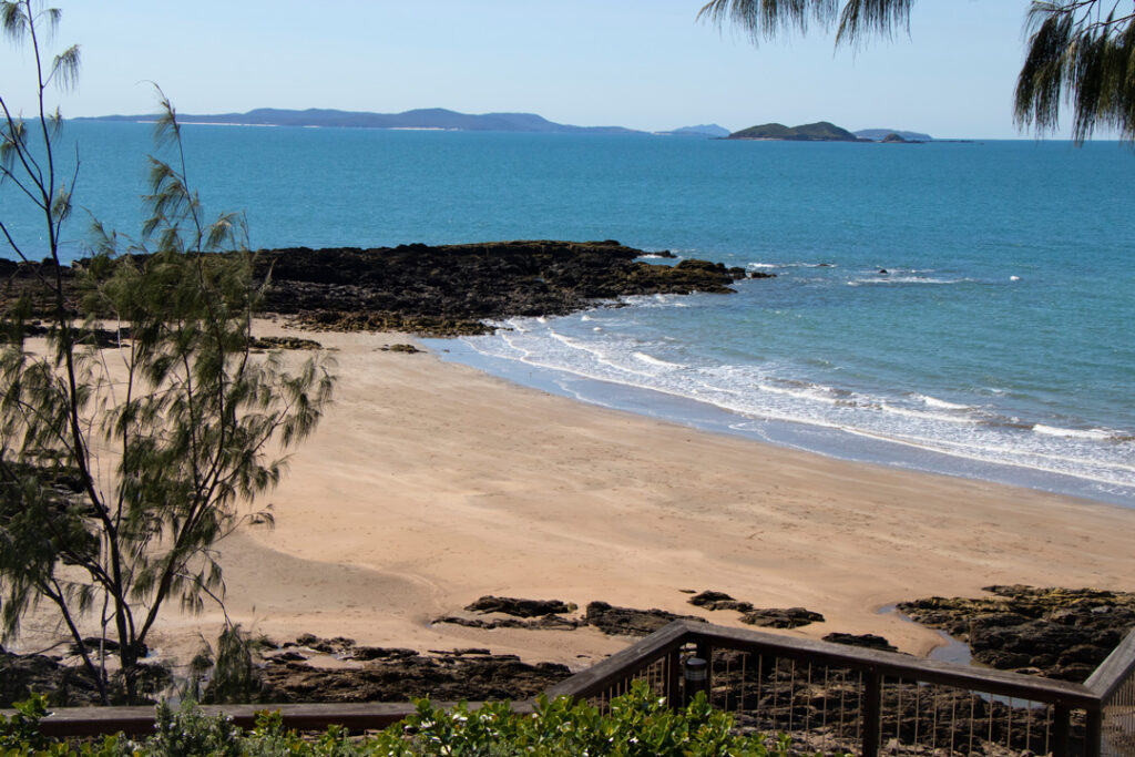 A wooden deck onto a sandy beach cove flanked by dark rocks, leading out to a blue ocean with small islands visible on the horizon.