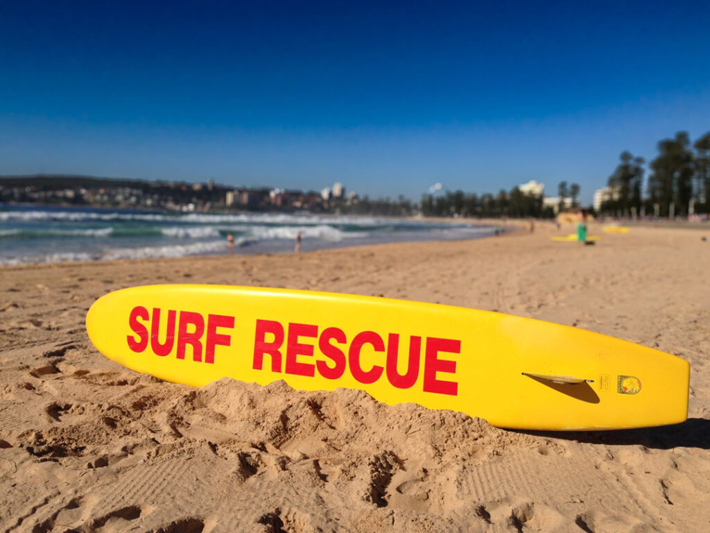 A yellow surf rescue board with bold red text resting on a sandy beach, with ocean waves and a coastline in the blurred background.