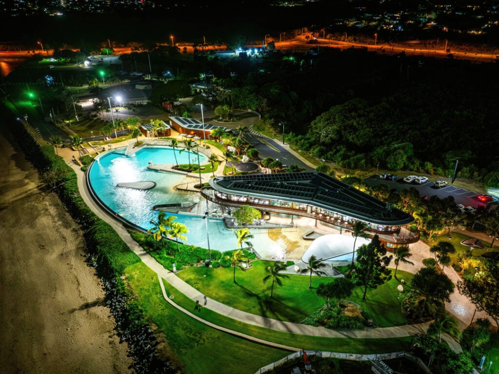 A modern lagoon-style public swimming pool complex illuminated by bright lights, situated along a dark beach coastline with a surrounding grassy park and parking area.